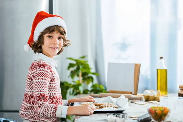 Side view of cute happy boy in santa hat holding baking tray with ginger cookies and smiling at camera — Stock Photo