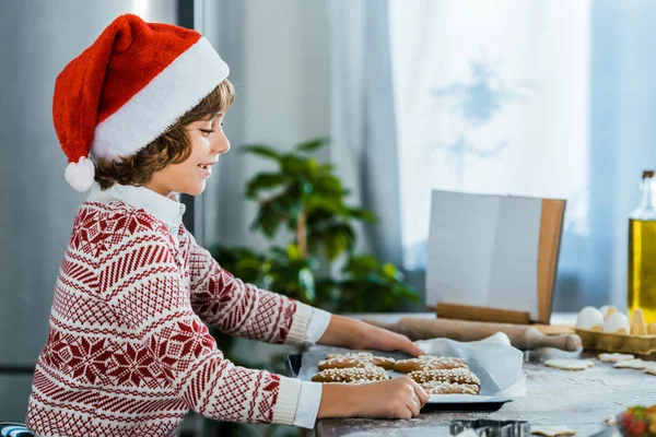 Vue latérale du garçon heureux dans le chapeau de Père Noël regardant la plaque de cuisson avec des biscuits au gingembre — Photo de stock