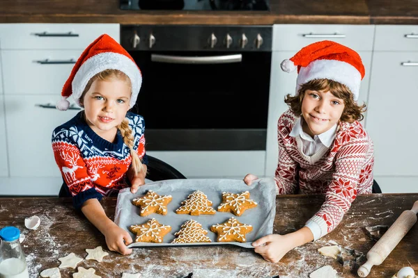 Vista de ángulo alto de lindos niños felices en sombreros de santa celebración bandeja para hornear con galletas de jengibre y sonriendo a la cámara - foto de stock