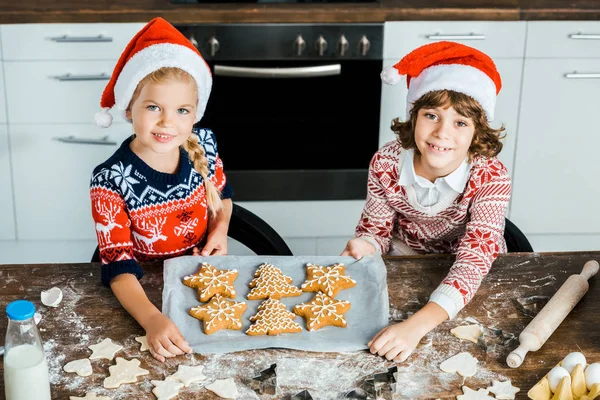 Blick aus der Vogelperspektive auf entzückende glückliche Kinder in Weihnachtsmützen, die ein Blech mit Ingwerplätzchen halten und in die Kamera lächeln — Stockfoto