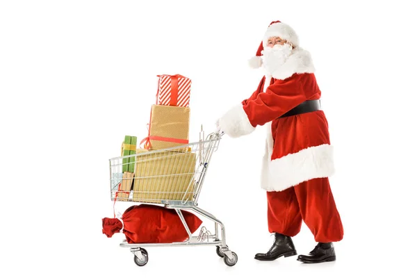 Vista lateral de Santa Claus caminando con carrito de compras lleno de cajas de regalo aisladas en blanco - foto de stock