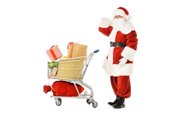 Visão lateral de Papai Noel perplexo com carrinho de compras olhando para a câmera isolada no branco — Fotografia de Stock