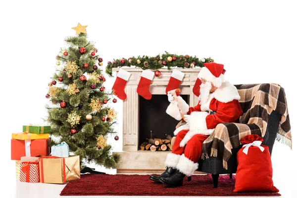 Santa claus con carta sentado en sillón cerca de la chimenea y el árbol de Navidad aislado en blanco - foto de stock