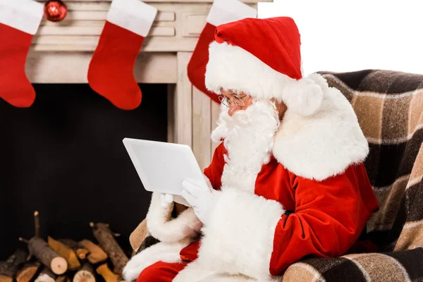 Enfocado santa claus usando tableta mientras está sentado en sillón cerca de la chimenea aislado en blanco - foto de stock