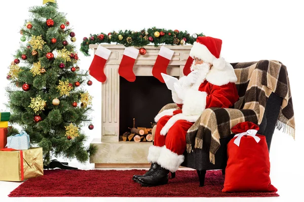 Père Noël en utilisant la tablette tout en étant assis dans le fauteuil près de la cheminée isolé sur blanc — Photo de stock
