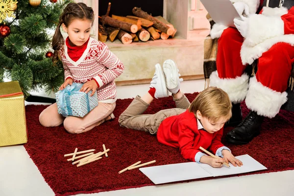 Recortado disparo de los niños desembalaje regalos y dibujo en el suelo delante de santa - foto de stock