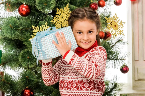 Niño sonriente sosteniendo regalo de navidad delante del árbol de navidad - foto de stock