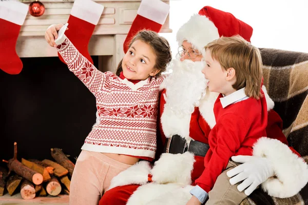 Glückliche Kinder machen Selfie mit dem Weihnachtsmann, während sie zusammen im Sessel sitzen — Stockfoto
