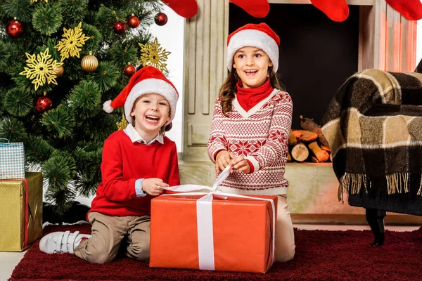 Happy little kids opening red christmas gift while sitting on floor — Stock Photo