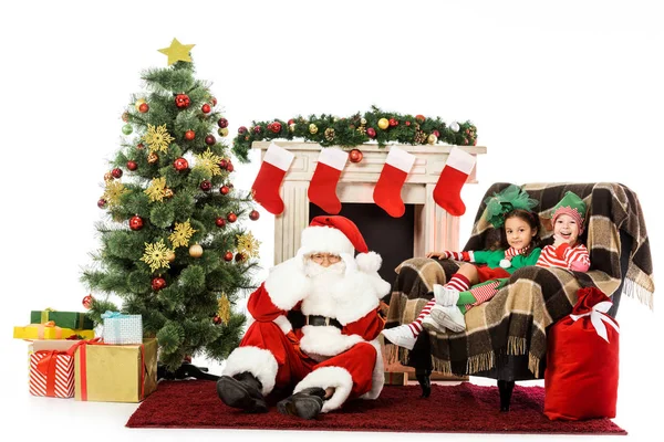 Niños y santa sentado cerca de la chimenea y el árbol de Navidad y mirando a la cámara aislada en blanco - foto de stock