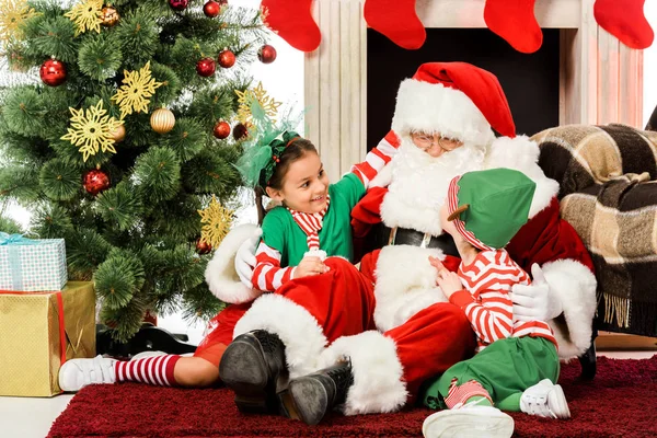 Babbo Natale abbraccia i bambini eccitati mentre si siedono sul pavimento davanti al camino insieme — Foto stock