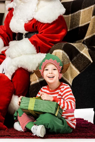 Niño feliz sentado en la alfombra con regalo de Navidad cerca de santa en sillón - foto de stock