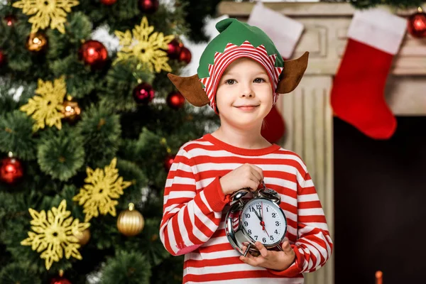 Adorável criança em traje de duende com despertador de pé na frente da árvore de natal — Stock Photo