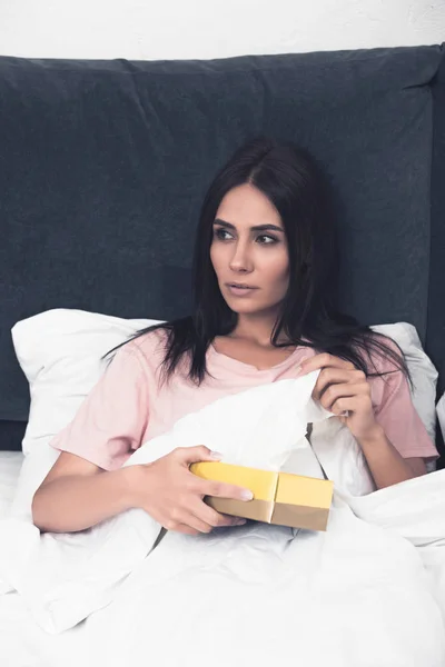 Mujer joven enferma tomando servilleta de papel fuera de la caja mientras está sentado en la cama - foto de stock