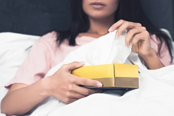 Recortado disparo de enfermo joven mujer tomando papel servilleta fuera de caja mientras está sentado en la cama - foto de stock