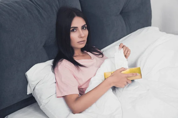 Malade jeune femme avec boîte de serviettes en papier assis dans le lit et regardant la caméra — Photo de stock