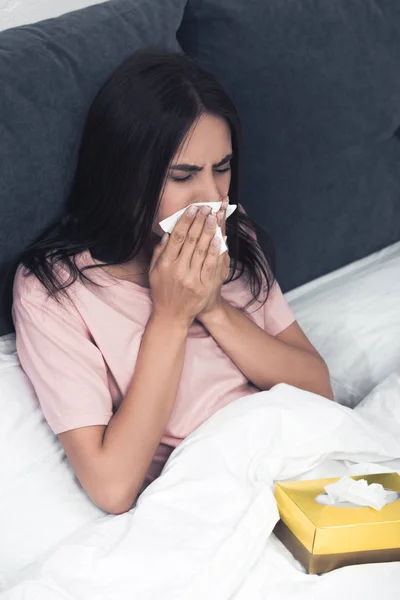 Mujer joven enferma con caja de servilletas de papel estornudando mientras está sentada en la cama - foto de stock