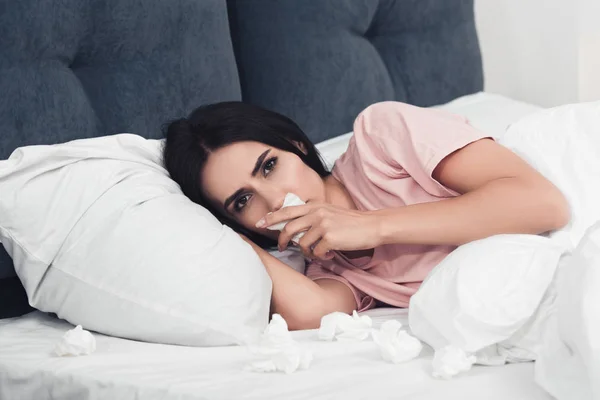 Close-up shot of sick young woman with runny nose lying in bed with napkins — Stock Photo