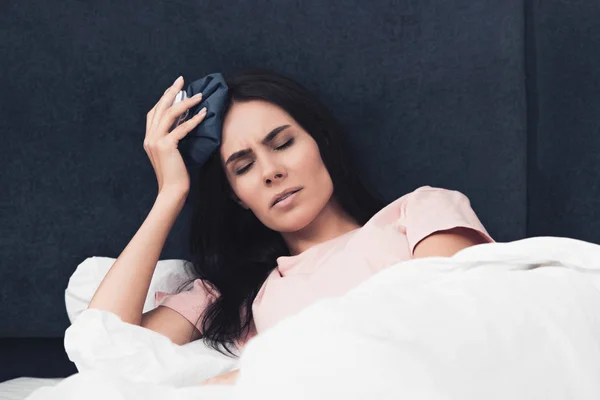 Suffering sick young woman holding ice pack on head while lying in bed — Stock Photo