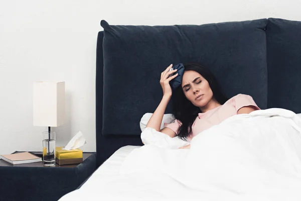 Страждаюча хвора молода жінка тримає лід на голові, лежачи в ліжку — стокове фото