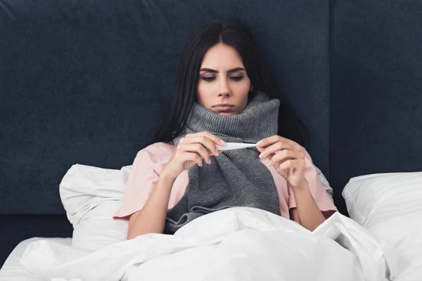 Mujer joven enferma en bufanda mirando el termómetro electrónico mientras está acostada en la cama — Stock Photo