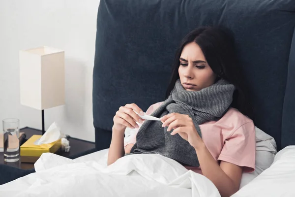 Mujer joven enferma en bufanda mirando el termómetro eléctrico mientras está acostada en la cama - foto de stock