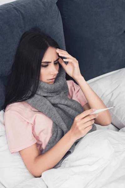 Хвора молода жінка в шарфі дивиться на електронний термометр, лежачи в ліжку — стокове фото