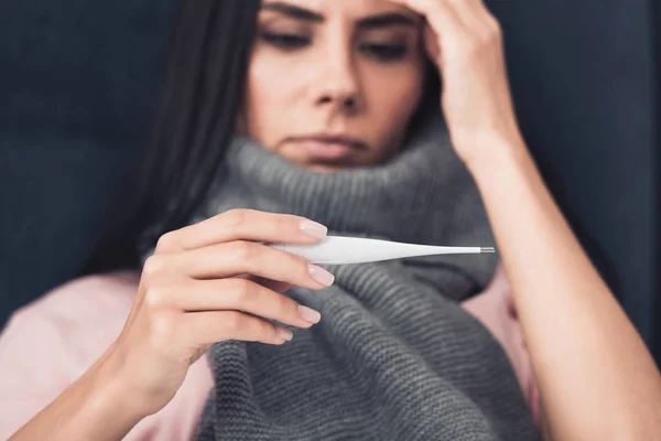 Primer plano de mujer joven enferma en bufanda mirando termómetro electrónico - foto de stock