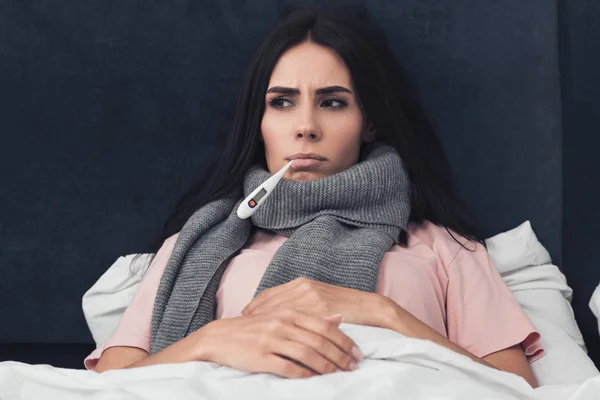 Jeune femme malade mesurant la température avec thermomètre buccal alors qu'elle est couchée au lit — Photo de stock
