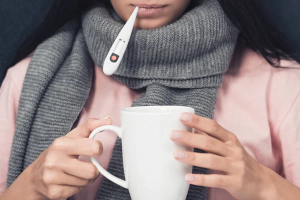 Tiro recortado de doente jovem mulher medir a temperatura com termômetro boca e segurando xícara de bebida quente — Fotografia de Stock