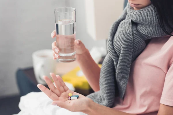 Cropped shot of sick young woman holding pills and glass of water while lying in bed — Stock Photo