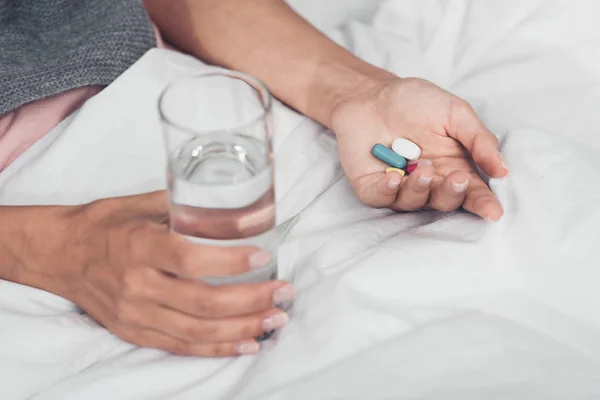 Inyección recortada de mujer enferma sosteniendo píldoras y vaso de agua mientras está acostado en la cama - foto de stock
