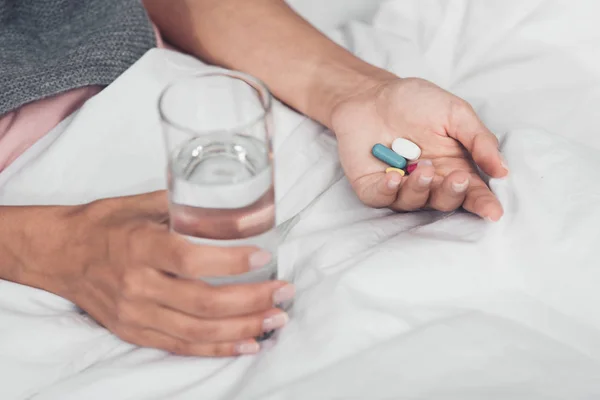 Cropped shot of sick woman holding pills and glass of water while lying in bed — Stock Photo
