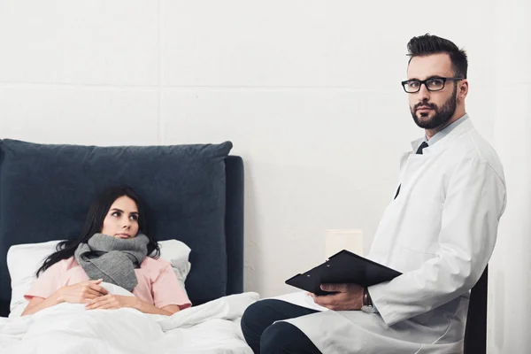 Schöner junger Arzt sitzt mit Klemmbrett und schaut in die Kamera, während Patientin im Bett liegt — Stockfoto