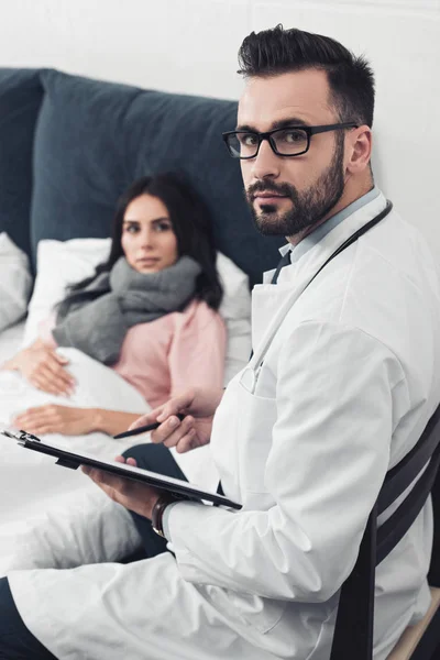 Guapo joven médico sentado con portapapeles y mirando a la cámara mientras paciente mujer acostada en la cama en el fondo - foto de stock
