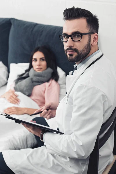 Beau jeune médecin assis avec presse-papiers et regardant la caméra tandis que la patiente couchée au lit sur le fond — Photo de stock