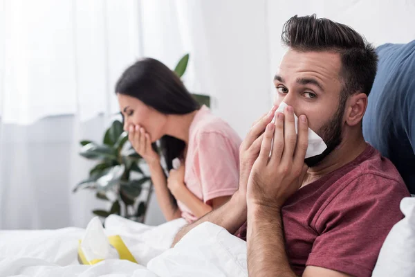 Diseased young sick couple sneezing into paper napkins while sitting in bed — Stock Photo