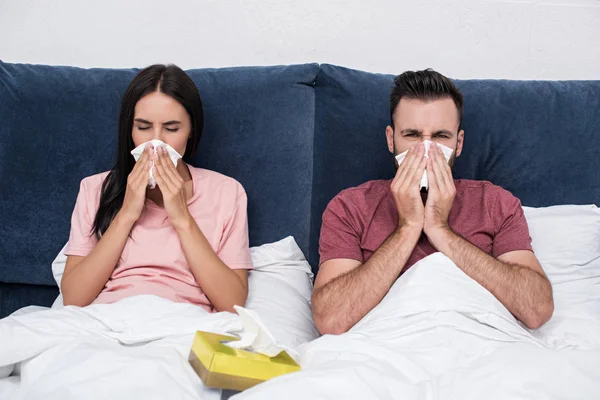 Joven pareja enferma estornudando en servilletas de papel mientras está sentado en la cama - foto de stock