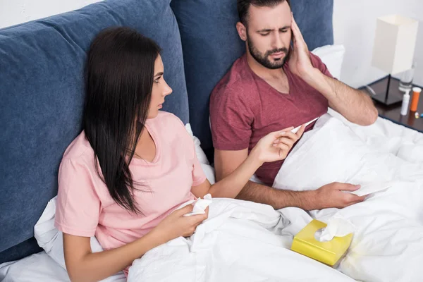 Young woman passing electric thermometer to her sick boyfriend in bed — Stock Photo