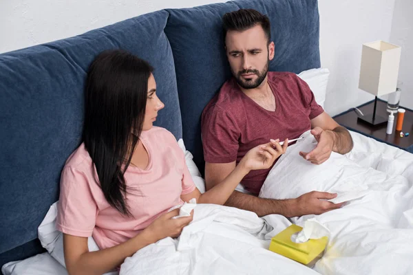 Mujer pasando termómetro eléctrico a su novio enfermo en la cama — Stock Photo