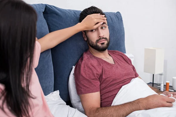 Cropped shot of young woman touching forehead of sick husband while sitting in bed — Stock Photo
