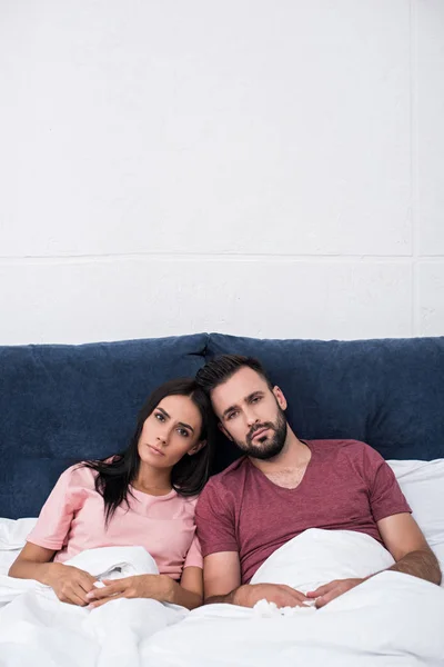 Serious young couple leaning at each other while sitting in bed and looking at camera — Stock Photo