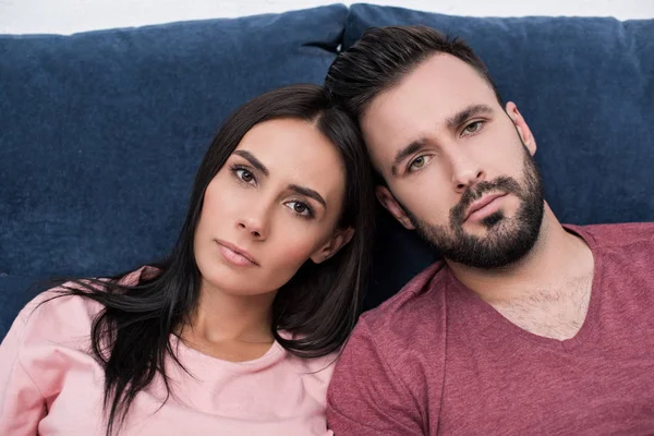 Unemotional young couple leaning at each other while sitting on couch and looking at camera — Stock Photo