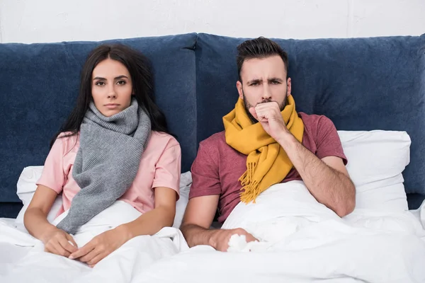 Enfermo joven pareja con tos sentado en la cama y mirando a la cámara - foto de stock
