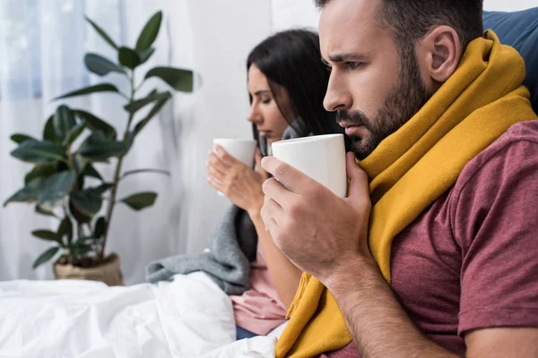 Sick young couple in scarves sitting in bed with cups of hot drink — Stock Photo