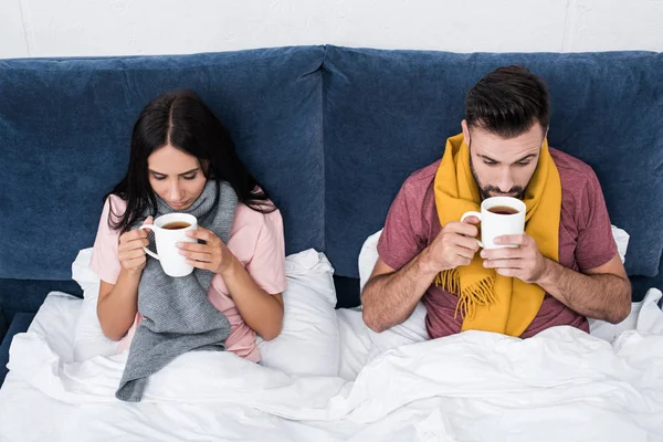 Vue grand angle de jeune couple malade dans des écharpes assis dans le lit avec des tasses de boisson chaude — Photo de stock