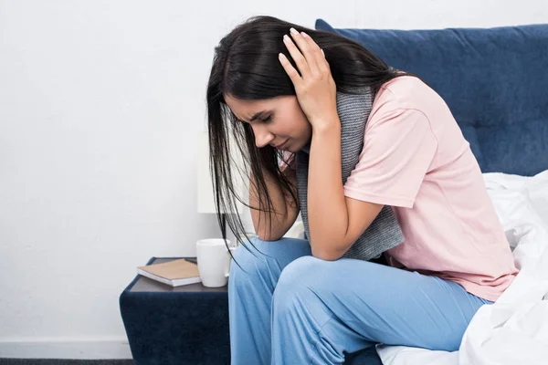 Junge Frau mit Schal leidet unter Kopfschmerzen, während sie im Bett sitzt — Stockfoto