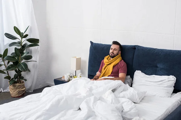 Подавленный больной молодой человек лежит в постели в шарфе и смотрит в сторону — стоковое фото