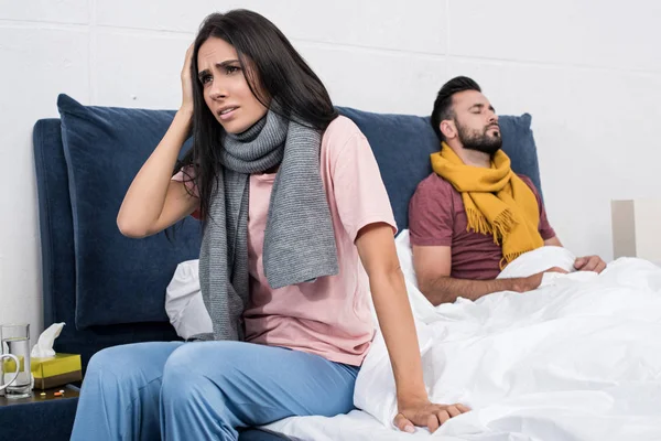Больная молодая пара в шарфах, сидящая на кровати вместе — стоковое фото