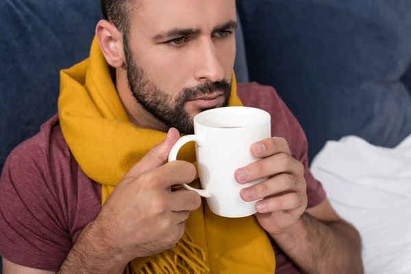 Malade jeune homme avec écharpe tenant tasse de thé chaud au lit — Photo de stock